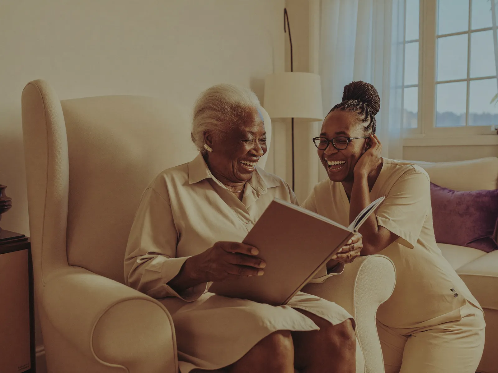 A senior resident and caregiver share a laugh over a photo album in a bright, cozy living room.
